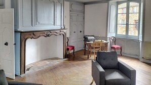 A room with a fireplace, a wooden table with chairs, a window with a view of buildings, and a radiator. - L' Arche d' Yvann (Campuac)