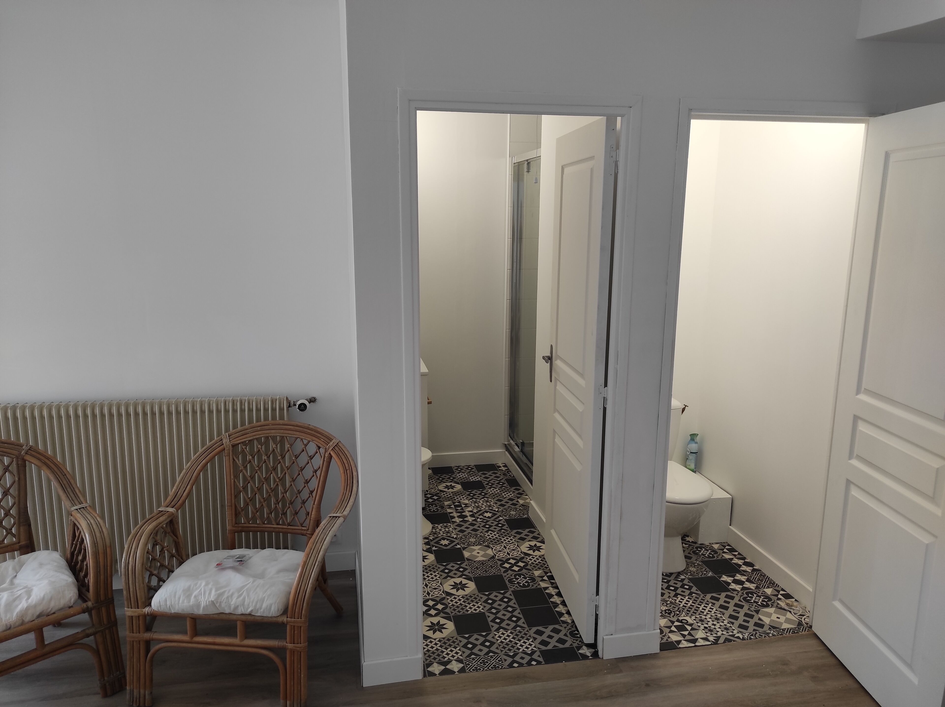 Una habitación con un radiador, dos sillas de mimbre, un suelo estampado y una puerta que da a un baño con inodoro y ducha.