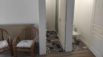 Una habitación con un radiador, dos sillas de mimbre, un suelo estampado y una puerta que da a un baño con inodoro y ducha.