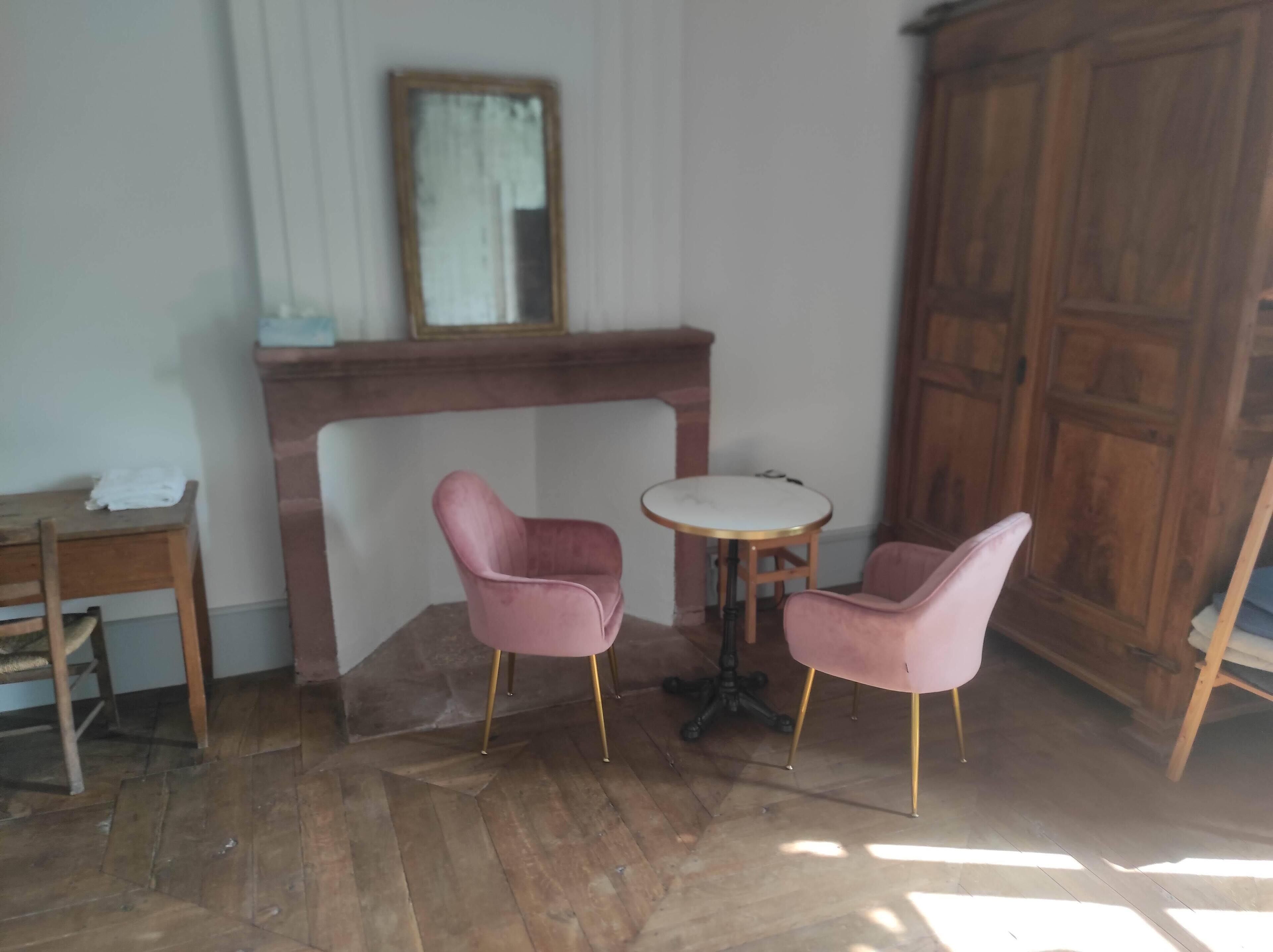 A room with a wooden floor, a fireplace, two pink chairs, a small round table, and a wooden cabinet.