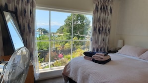 A bedroom with a large window, a bed with folded towels, a bedside table with a lamp, and a fan.