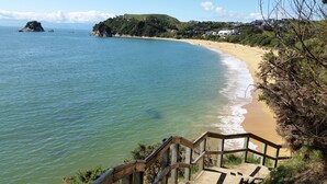 A coastal view with a sandy beach, a wooden staircase leading down to the water, and a hill with vegetation and buildings in the background.
