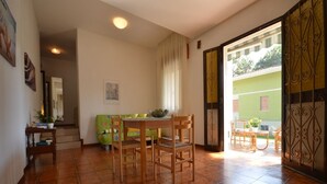 A dining area with a table and chairs, a green sofa, and a view of a green building through an open door. - Ville Missana (Lignano Sabbiadoro)