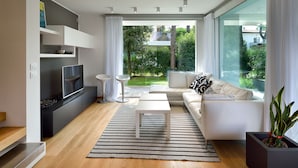 A modern living room with a TV, sofa, coffee table, and large windows offering a view of a garden. - Ville Summer (Lignano Sabbiadoro)