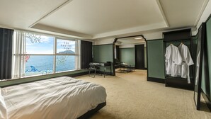 A hotel room with a bed, a desk, and a chair. There is a window with a view of the sea and a sign that says "YOU HAVING FUN?". There are clothes hanging on a rack. - Island.F Jeju (Seogwipo)