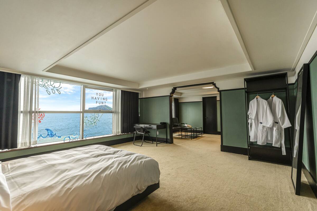 A hotel room with a bed, a desk, and a chair. There is a window with a view of the sea and a sign that says "YOU HAVING FUN?". There are clothes hanging on a rack.