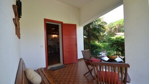 A wooden bench and a chair on a balcony with a red door, a small table with a plant, and a view of greenery. - Villedda (Lignano Sabbiadoro)