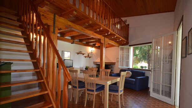 A wooden staircase leading to a lofted area with a dining table and chairs, a sofa, and a large window with a view of greenery. - Villedda (Lignano Sabbiadoro)