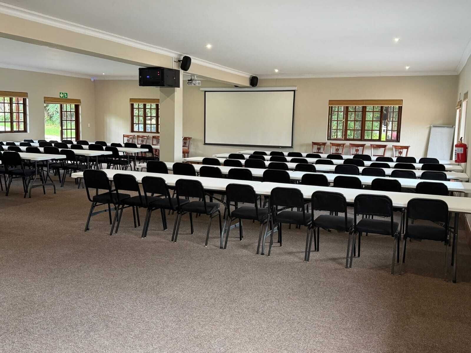 Een congreszaal met rijen tafels en stoelen, een beamer en een whiteboard.