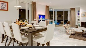 A modern dining area with a dark wooden table and white chairs, a television in the background, and a glass door leading to a balcony.