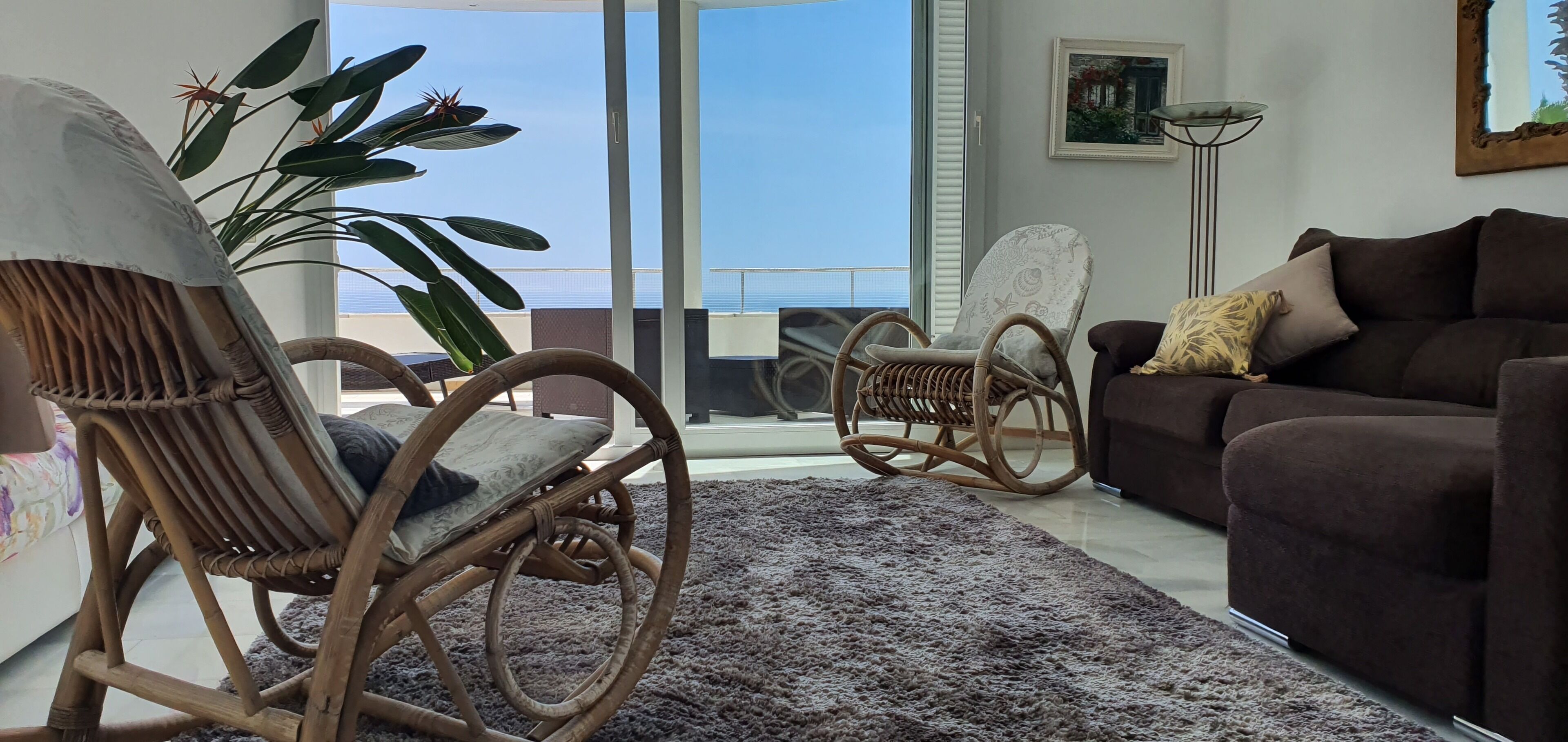 Una sala de estar con un sofá marrón, un sillón de mimbre, una planta grande en maceta y vista al mar a través de amplios ventanales.