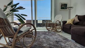Una sala de estar con un sofá marrón, un sillón de mimbre, una planta grande en maceta y vista al mar a través de amplios ventanales.