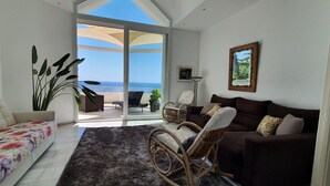 A spacious living area with a sofa, a brown couch, and a wicker chair. A large window offers a view of the ocean.