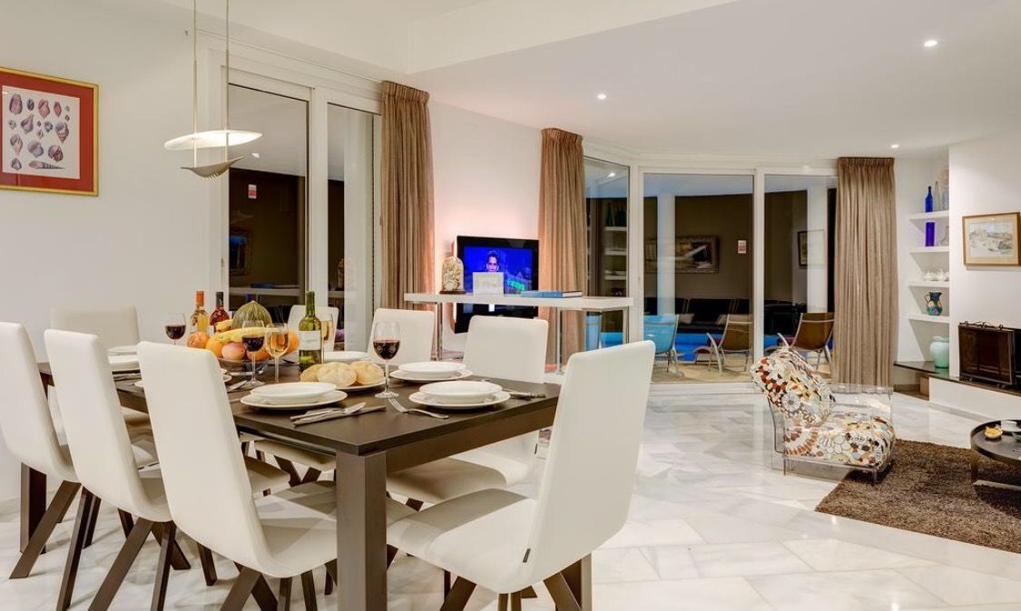 Un comedor moderno con una mesa de madera oscura y sillas blancas, un televisor al fondo y un vidrio que da a un balcón.