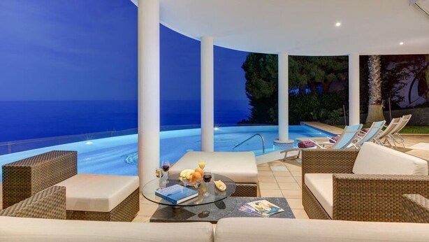 A luxurious outdoor lounge area with a view of a pool and the sea at dusk.