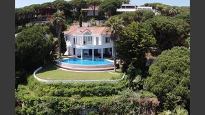 A large, white, two-story house with a circular pool surrounded by a low wall and lush greenery.