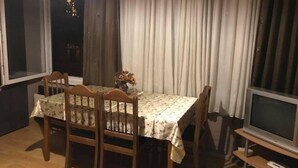 A dining area with a table covered by a floral tablecloth, surrounded by wooden chairs. A vase with flowers is on the table, and a television is on a stand to the right. - Apartment on Tsamebuli Ave (Tbilisi)