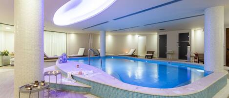 A modern indoor swimming pool with a curved edge, a sunken seating area, and a central table with candles.
