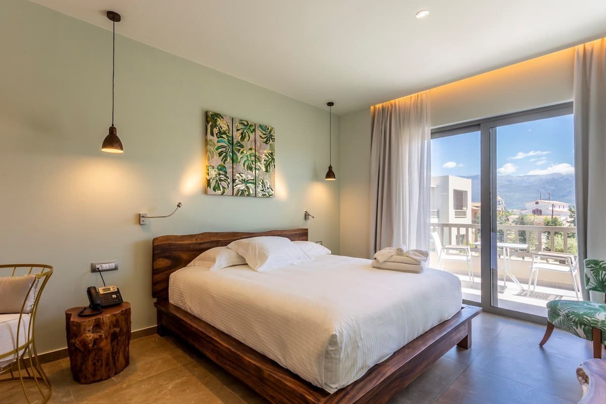 A bedroom with a large bed, a wooden headboard, a bedside table with a phone, and a view of the outside through a large window.