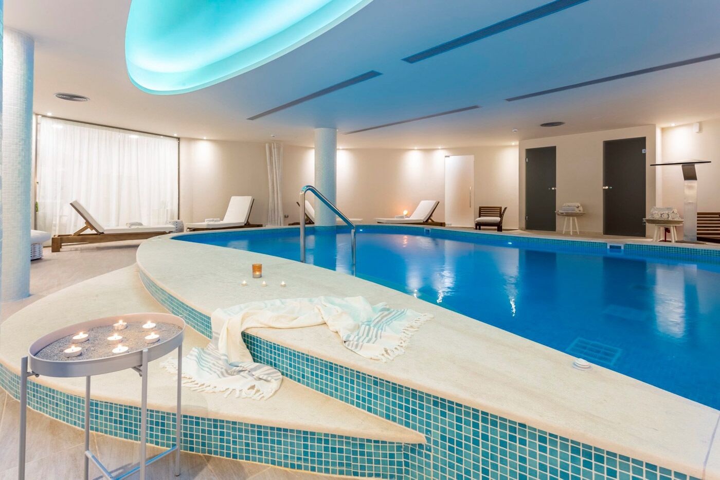 An indoor swimming pool with a tiled edge, lounge chairs, and a candle-lit area.