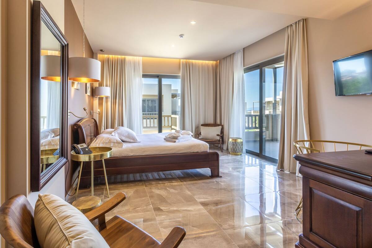 A hotel room with a large bed, a desk, a chair, a TV, and a balcony with curtains.