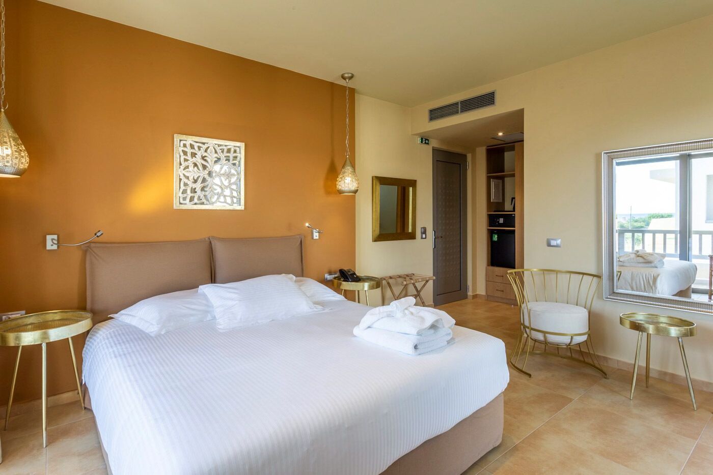 A hotel room with a large bed, a sitting area with a chair and a small table, a mirror, and a view of the outside balcony.