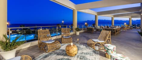 A rooftop terrace with wicker furniture, a round wooden table, and a view of the ocean at dusk.