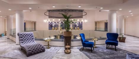 Een ruime lobby met een centraal zithoek, inclusief een gestoffeerde fauteuil, een blauwe fauteuil, en een ronde tafel met een potplant.