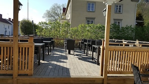 An outdoor patio with wooden deck, tables, and chairs, surrounded by greenery and buildings. - Gamleby Hotell (Gamleby)
