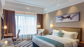Ein Hotelzimmer mit einem großen Bett, einem Schreibtisch mit Stuhl, einem kleinen Tisch und einem großen Fenster mit Vorhängen.