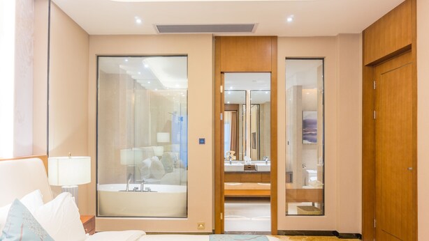 Une chambre d’hôtel comprenant un lit, une table de chevet, une lampe, un grand miroir, une armoire et une salle de bain visible à travers une porte en verre.