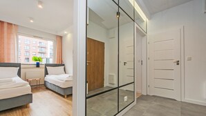 Une chambre d’hôtel moderne dotée d’un grand placard avec une porte coulissante en verre, d’un lit avec une tête de lit grise et d’une petite table sur laquelle est posée une plante.