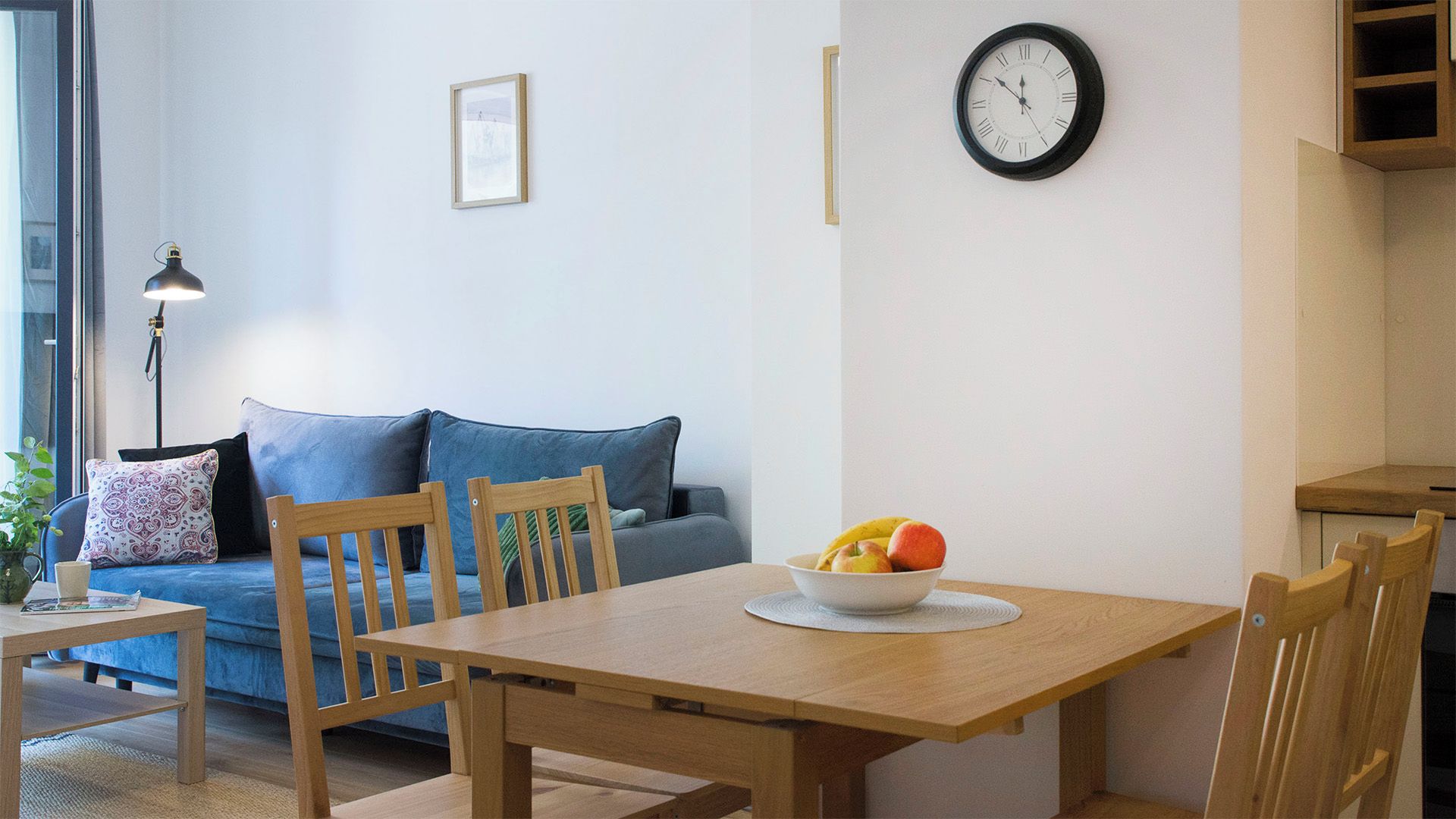A living room with a wooden dining table, chairs, a sofa, a clock, and a bowl of fruit.