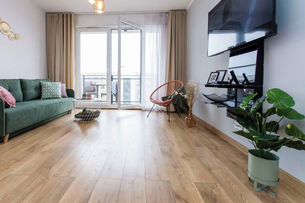 Een moderne woonkamer met een groene bank, een rode fauteuil, een aan de muur bevestigde televisie en een balkon met gordijnen.