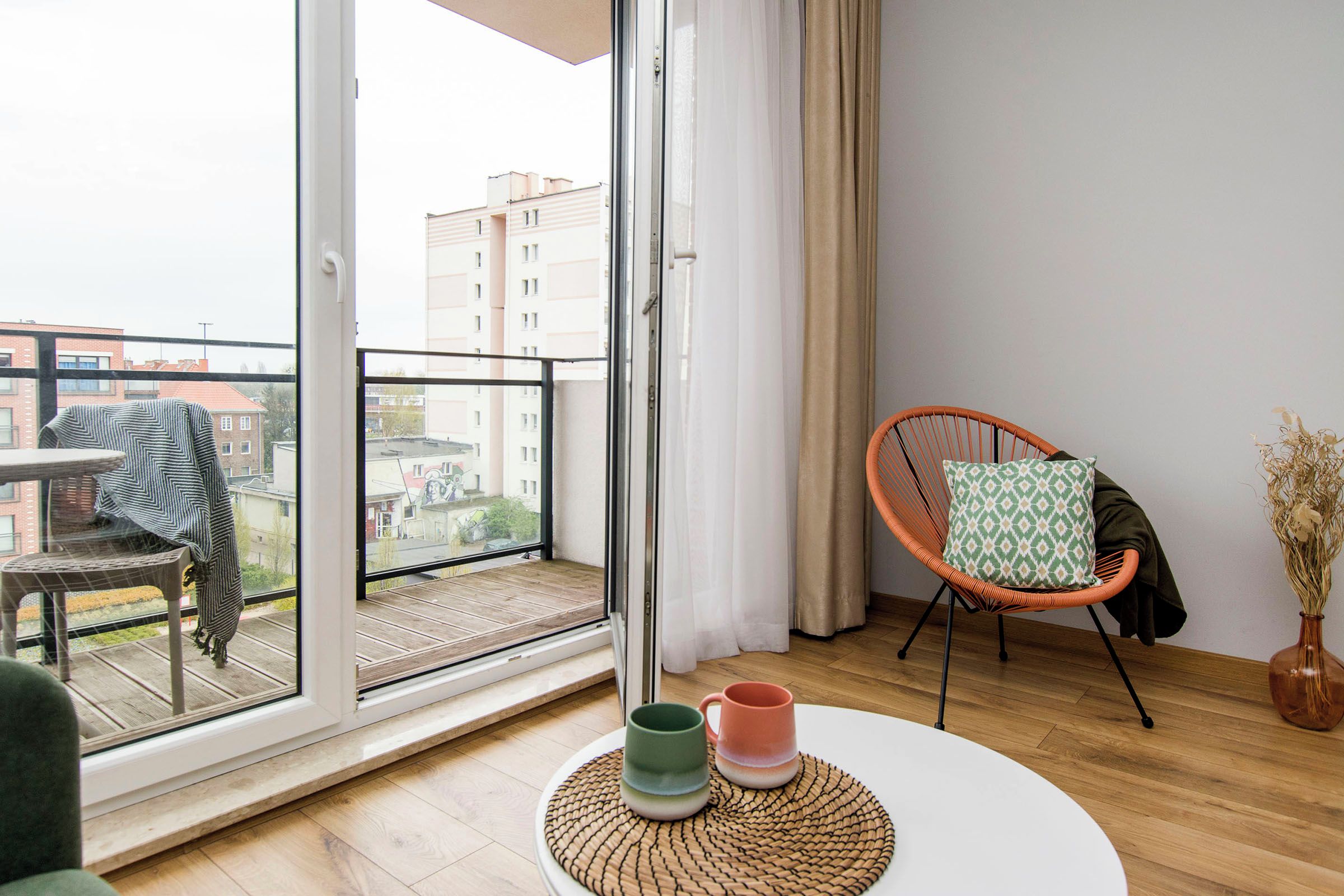 A modern living room with a balcony, a wicker chair, a round table with two mugs, and a vase with dried flowers.