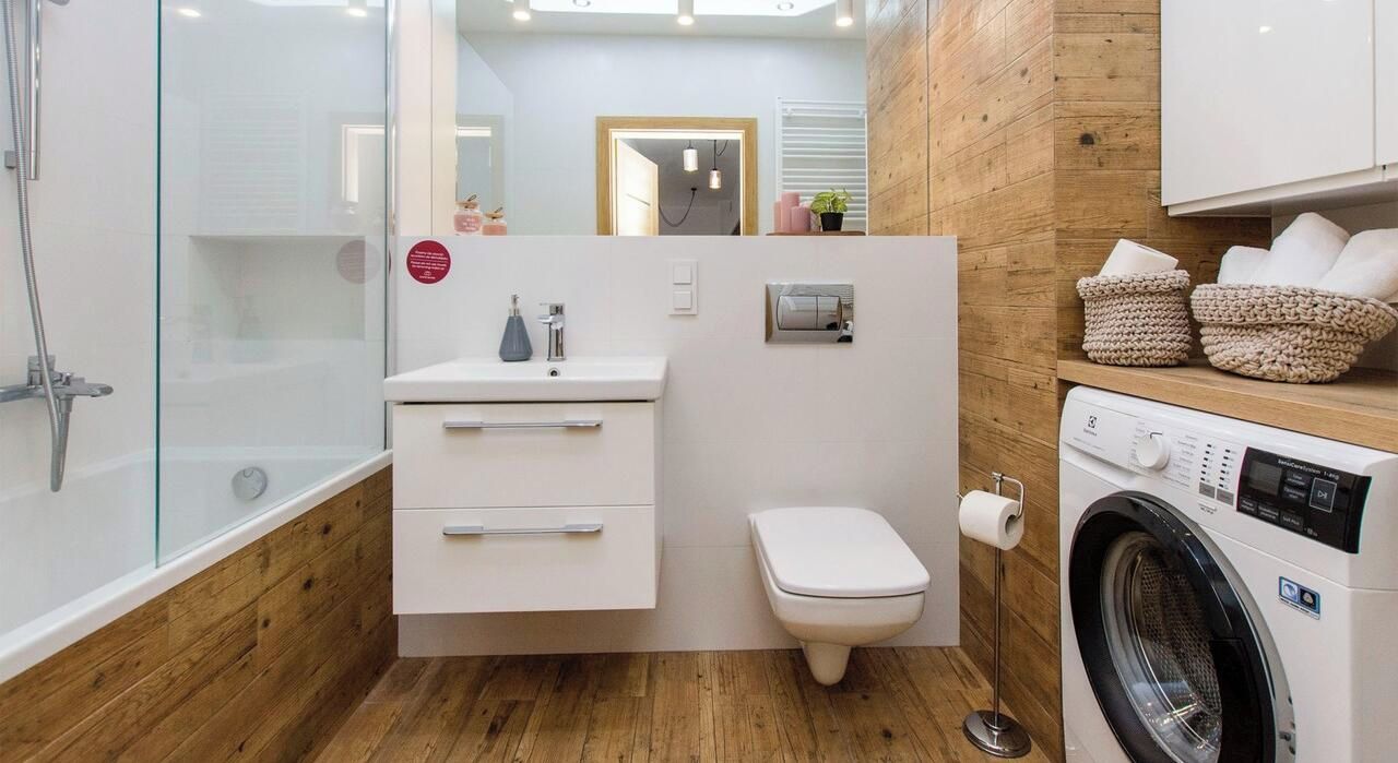 A compact bathroom with a glass shower enclosure, a white sink with a mirror, a toilet, and a washing machine.