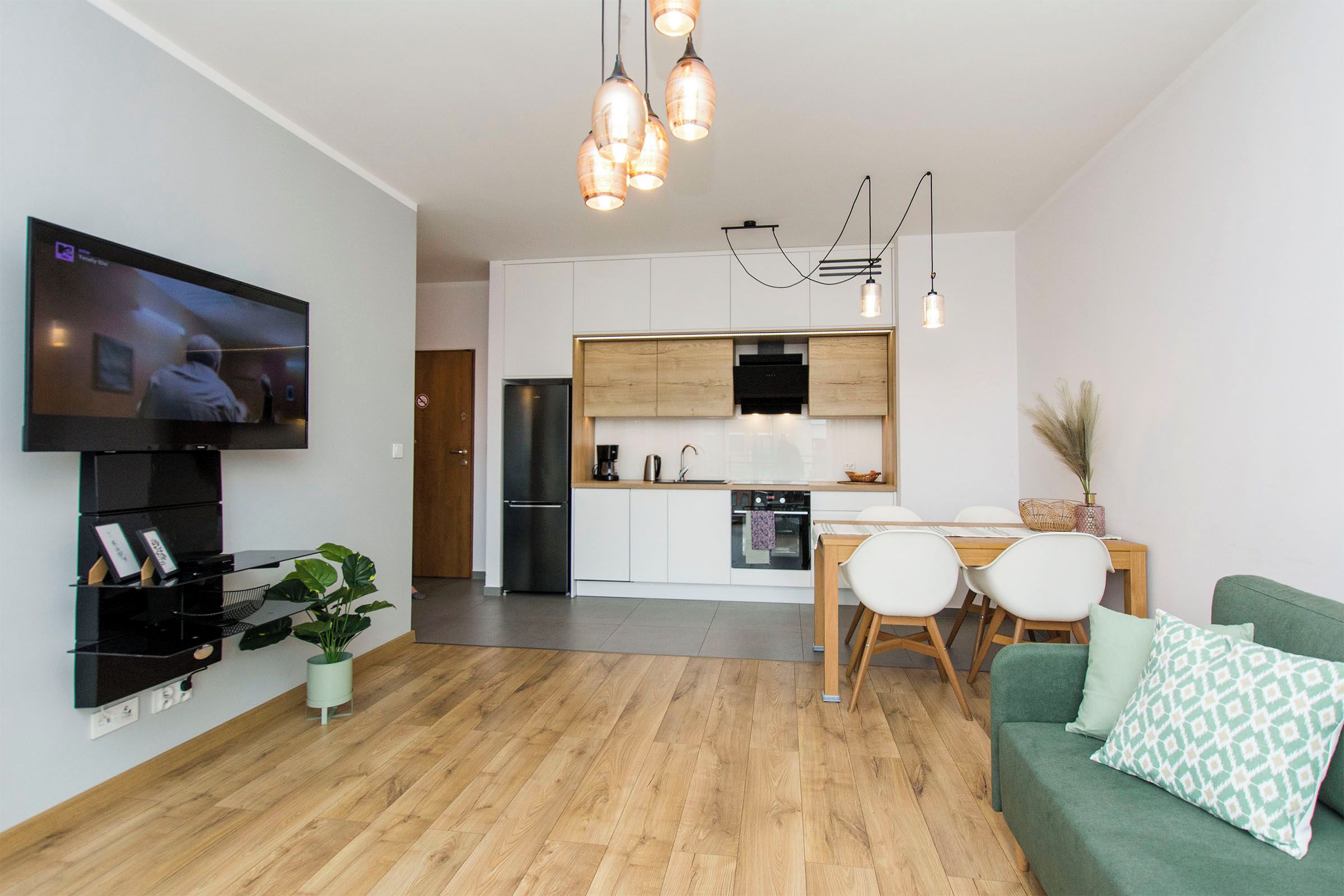 A modern living room with a flat-screen TV, a dining area with a wooden table and chairs, and a kitchen with white cabinets and a black refrigerator.