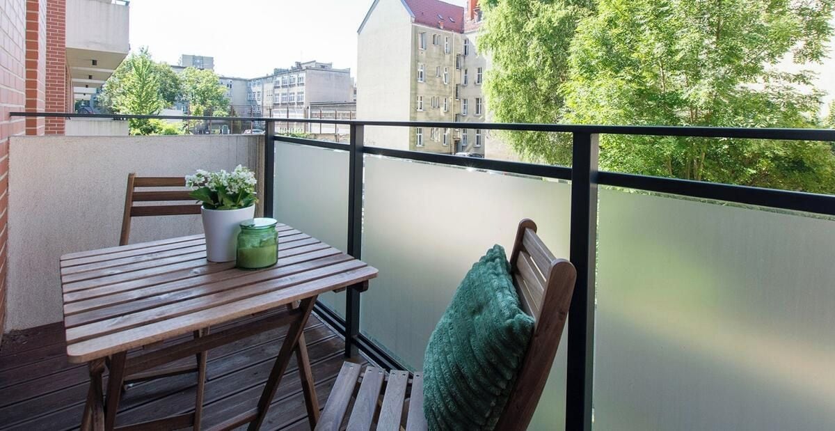 A balcony with a wooden table, chairs, a potted plant, and a view of buildings and trees.