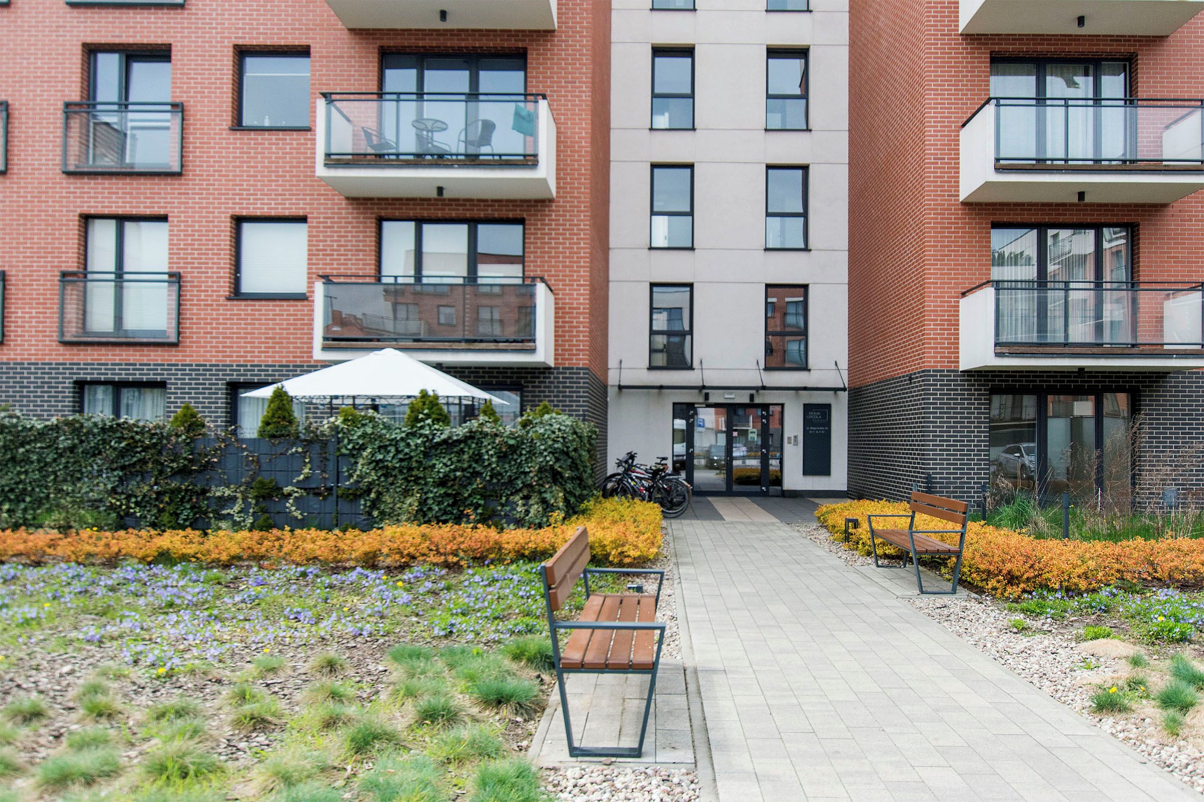 A multi-story residential building with balconies, a paved walkway, and a garden area with benches and flowers.