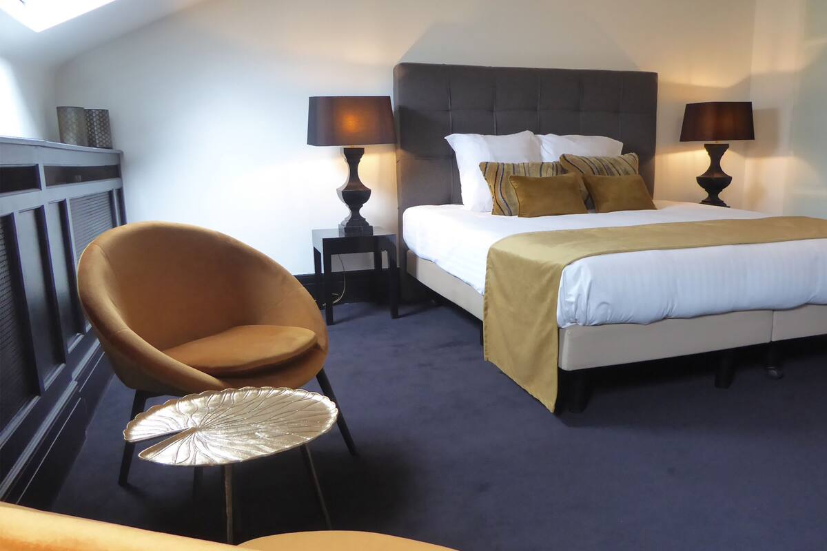 A hotel room with a bed, two bedside lamps, a chair, a small round table, and a view of the sky through a skylight.