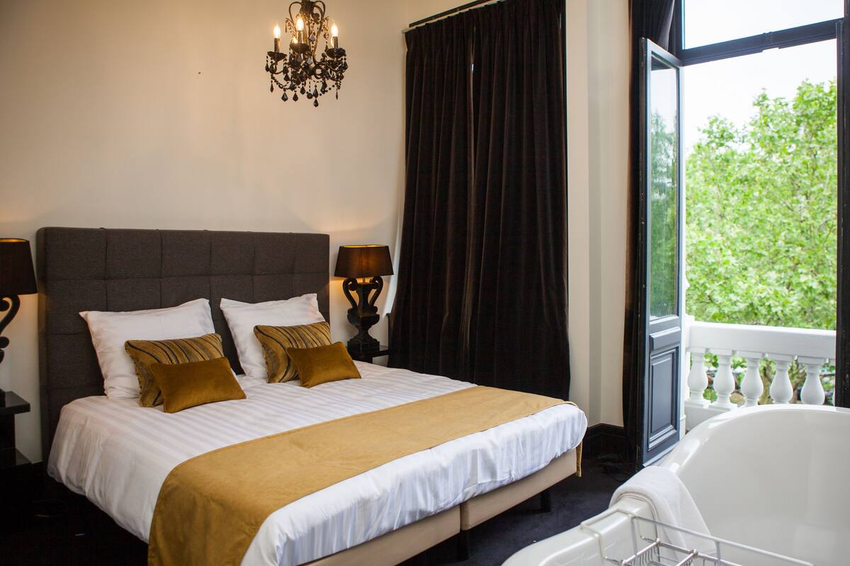 A bedroom with a large bed, two bedside lamps, a chandelier, a window with a view of greenery, and a white bathtub.