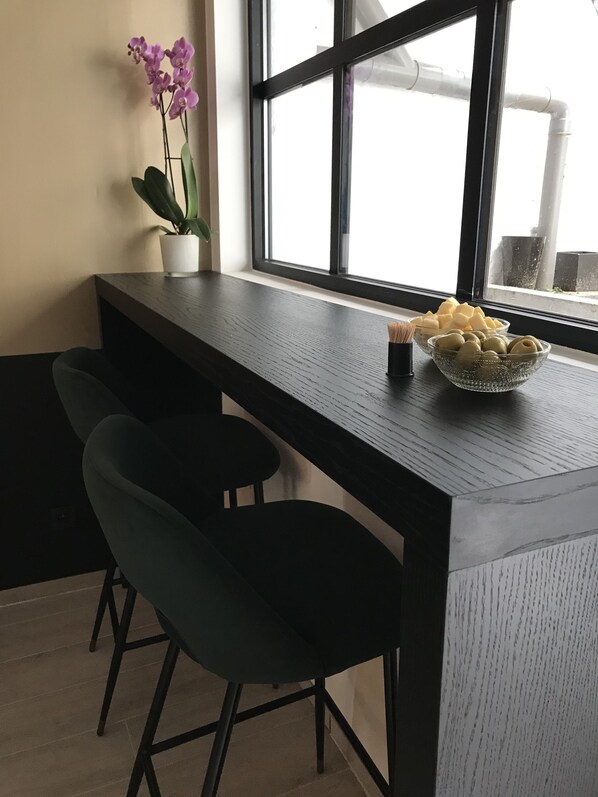 A modern kitchen with a dark wooden countertop, two bar stools, a potted orchid, and a bowl of food on the window sill.