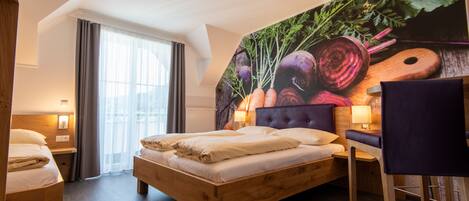 A modern hotel room with a wooden bed, a large fruit and vegetable mural, a sitting area with a chair, and a window with curtains.