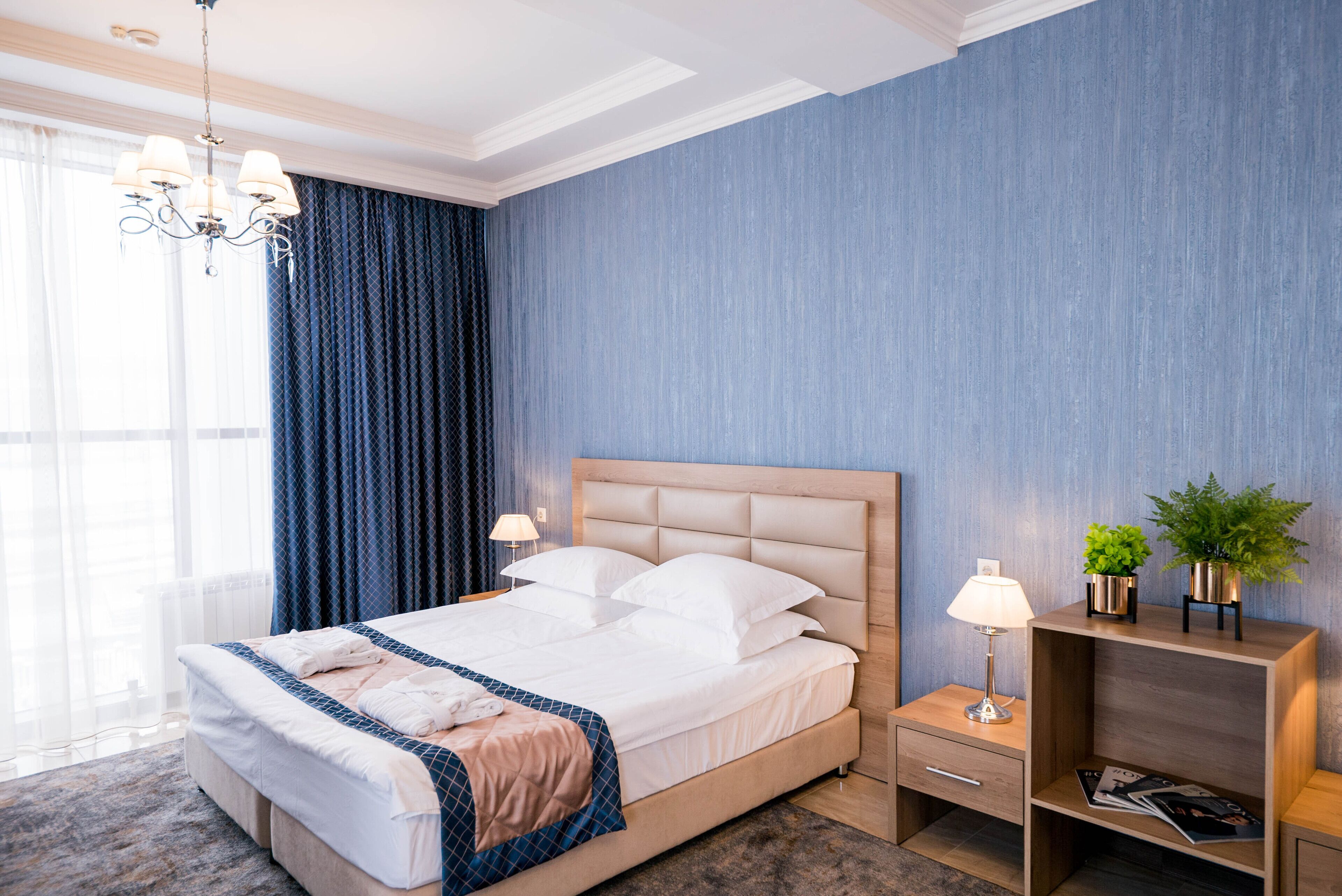 Ein modernes Hotelzimmer mit einem großen Bett, einem Nachttisch mit Lampe, einem kleinen Regal mit Pflanzen und Blick auf die Stadt durch große Fenster.