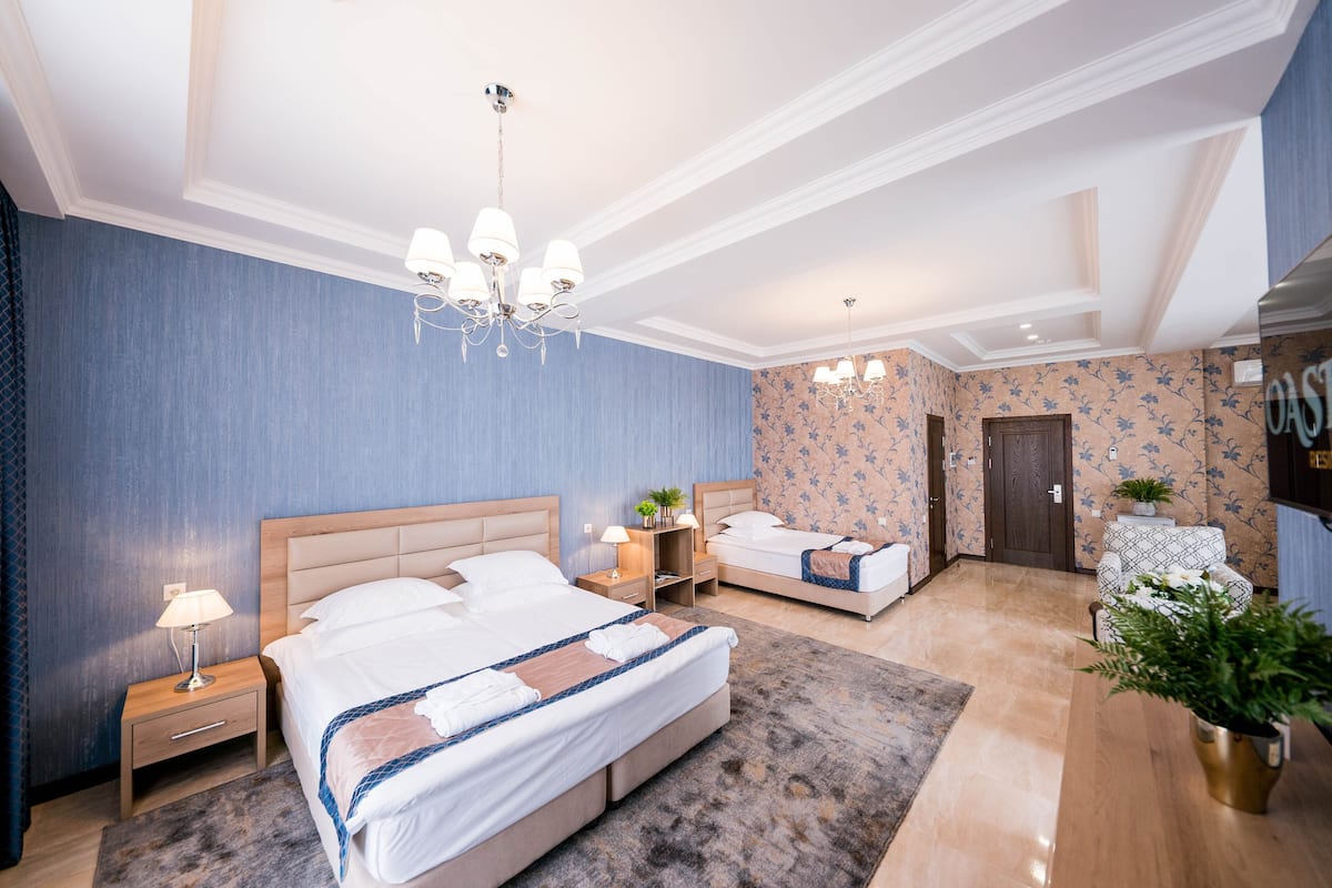 A hotel room with a large bed, two bedside tables with lamps, a small seating area with a chair, a potted plant, and a door leading to another room.