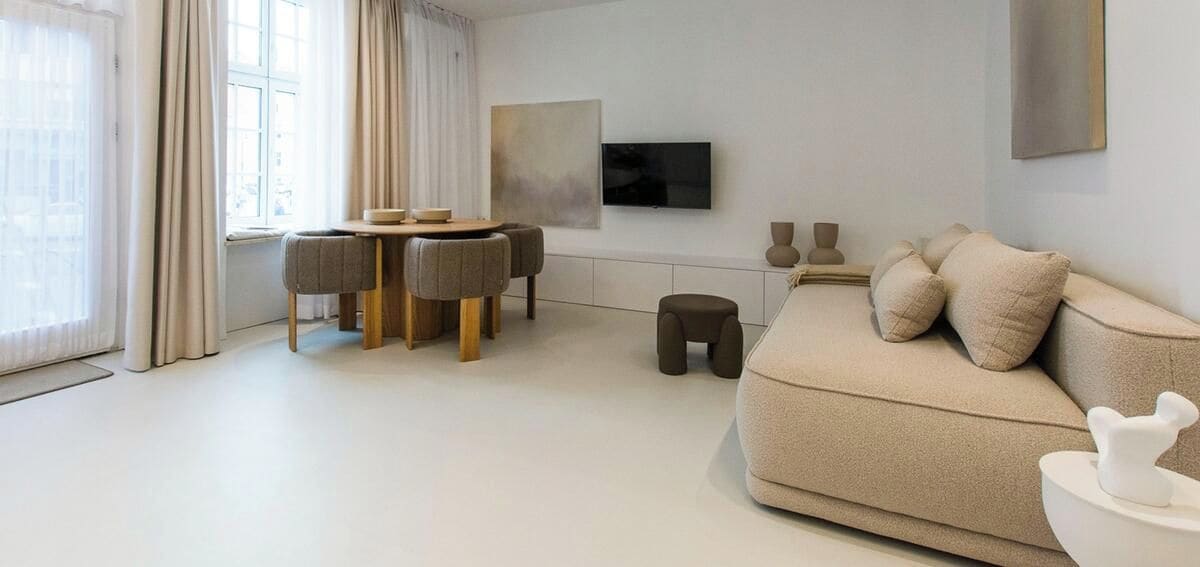 A modern living room with a sofa, a dining table with chairs, a TV mounted on the wall, and a window with curtains.