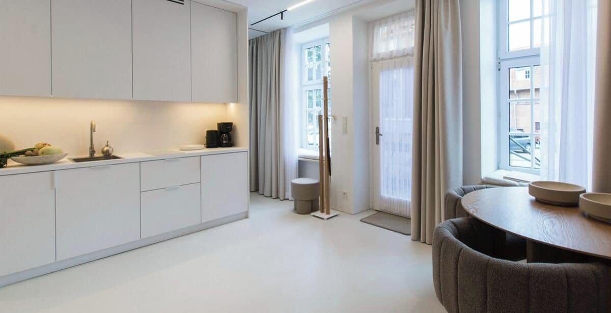 A modern kitchen with white cabinetry, a dining area with a round table, and a view of the street through large windows.