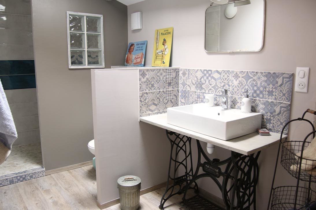 Un baño con un lavamanos blanco, un espejo y un salpicadero de azulejos. Hay una ventana, un inodoro y un cubo de basura.