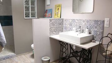 Un baño con un lavamanos blanco, un espejo y un salpicadero de azulejos. Hay una ventana, un inodoro y un cubo de basura.