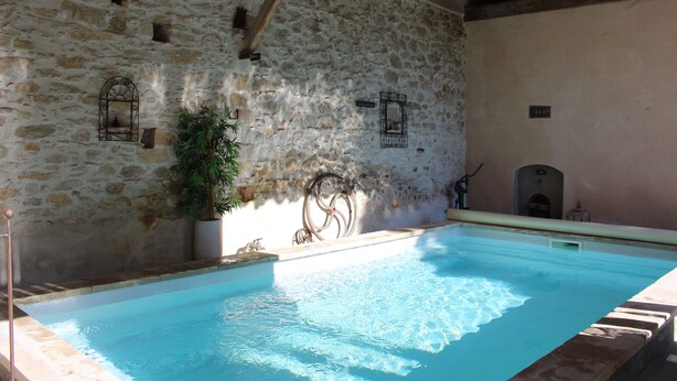 A stone-walled indoor swimming pool with a clear blue water surface, a potted plant, and a wooden ceiling beam.